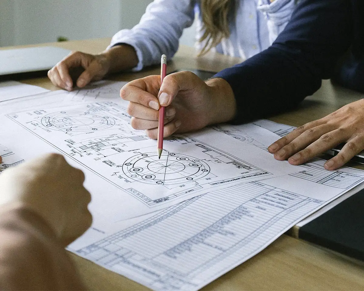 Ruke koje pišu i ukazuju na detaljne tehničke crteže, simbol saradnje i preciznosti u inženjerskom okruženju.