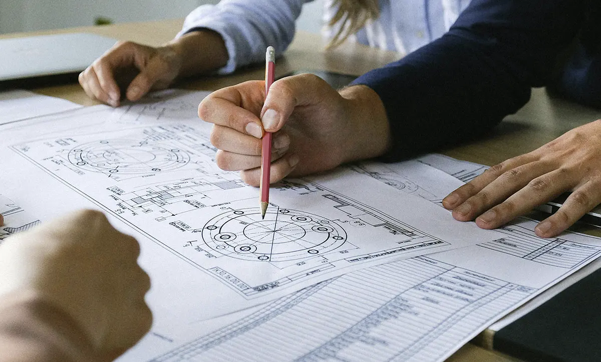 Ruke koje pišu i ukazuju na detaljne tehničke crteže, simbol saradnje i preciznosti u inženjerskom okruženju.