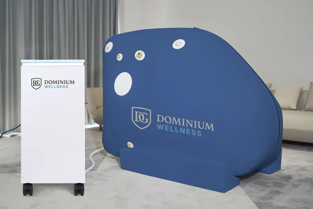 Plava hiperbarična komora Dominium Wellness, model DW160MS, postavljena pored belog uređaja u modernoj prostoriji sa zavesama i kaučem, u prijatnom i savremenom ambijentu.