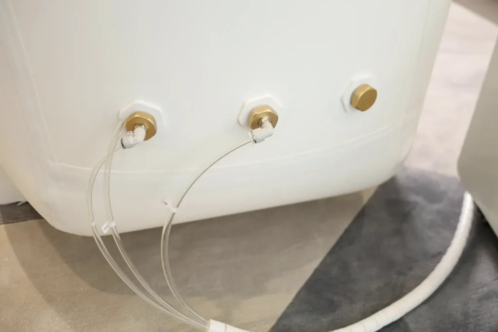 Detaljan prikaz zadnjeg dela bele hiperbarične komore, sa tri mesingana priključka i providnim crevima, u unutrašnjem prostoru sa svetlim podom.