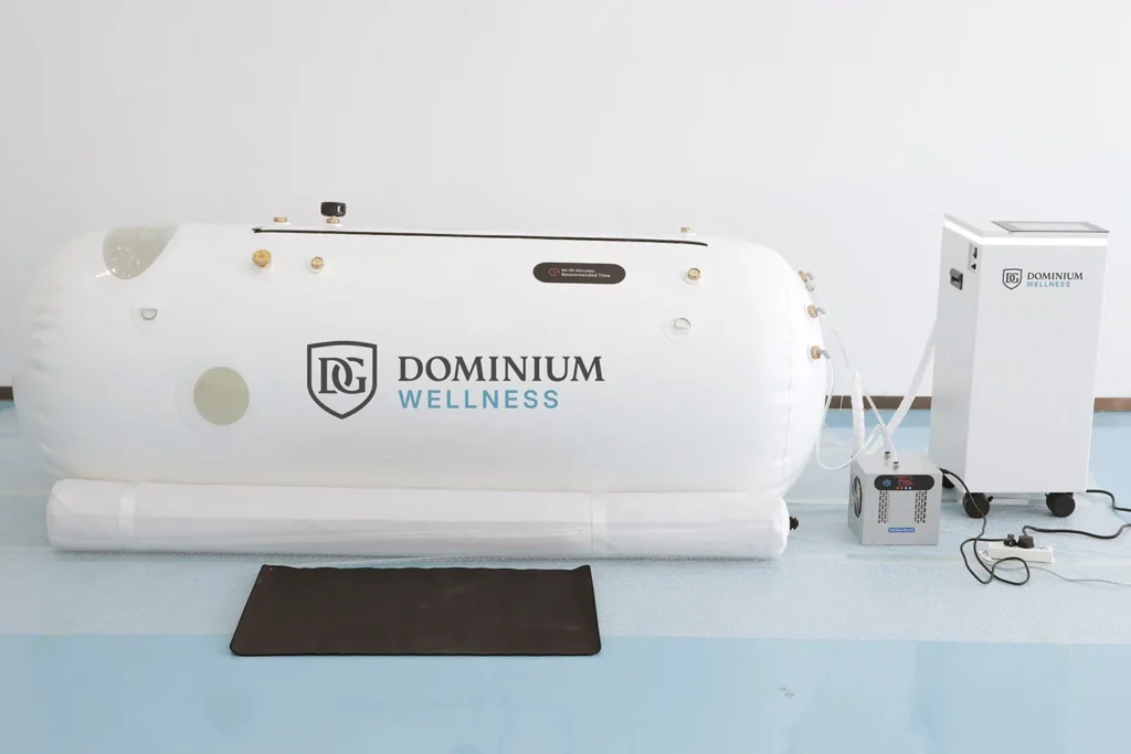 Naduvavajuća hiperbarična komora Dominium Wellness, model DW220ML, povezana sa kontrolnim jedinicama, postavljena na svetloplavom podu, u mirnom i kliničkom okruženju.