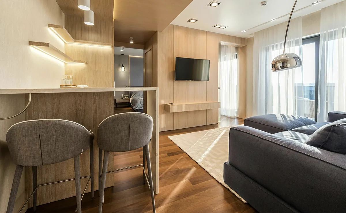 Moderan dnevni boravak sa elegantnim dizajnom, šankom sa dve sive barske stolice, drvenim detaljima, zidnim TV-om, velikom sivom garniturom i prozorima od poda do plafona.