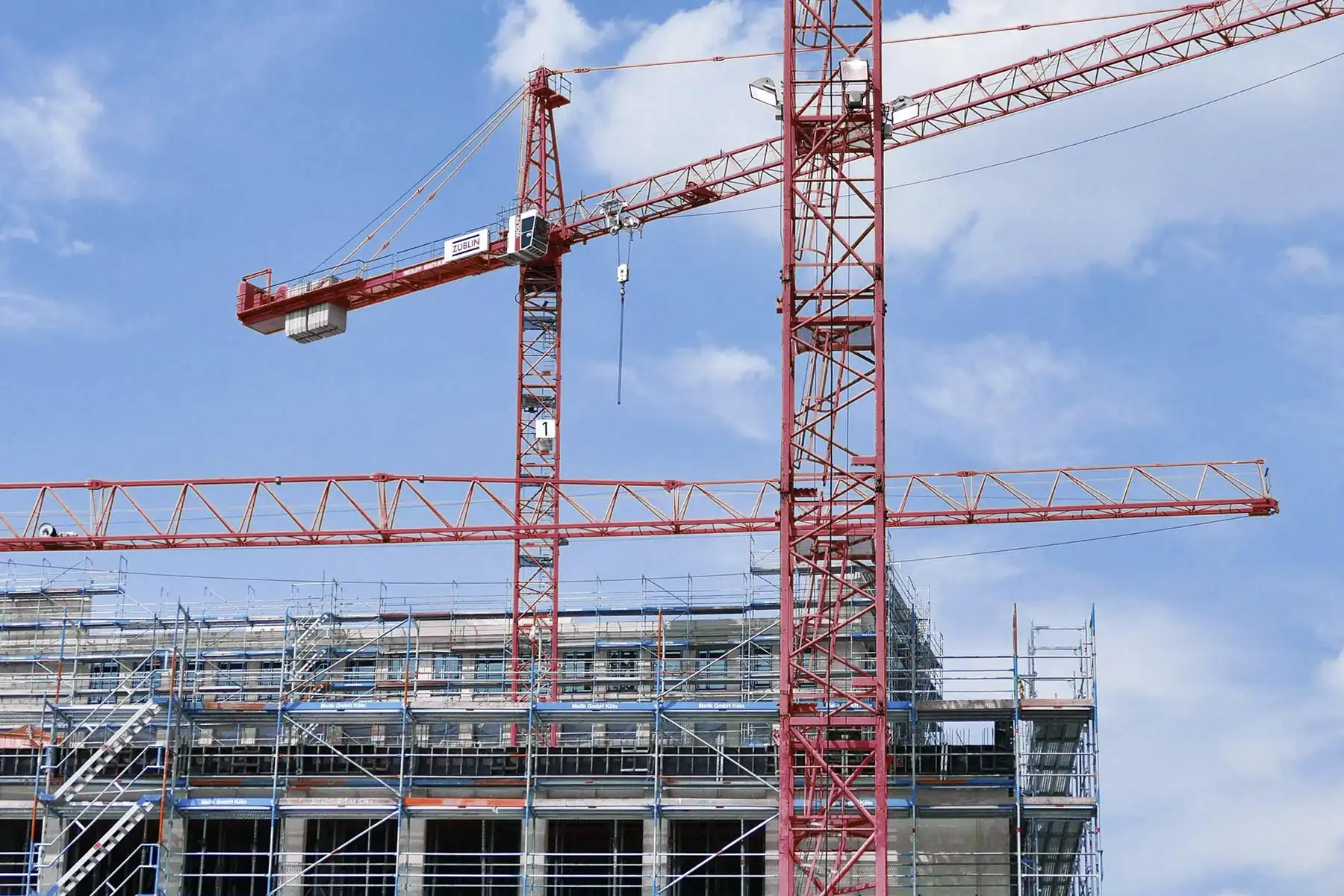 Crvene građevinske dizalice (kranovi) iznad objekta u izgradnji sa skelama, simbol rasta, razvoja i napretka.