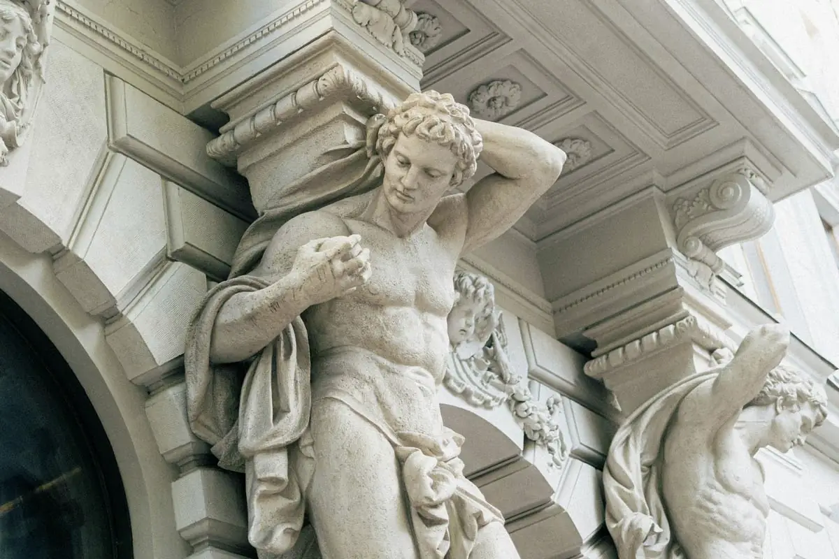 Kamena statua snažnog muškarca koji pridržava bogato ukrašenu fasadu zgrade, simbol snage i elegancije u arhitektonskom kontekstu.