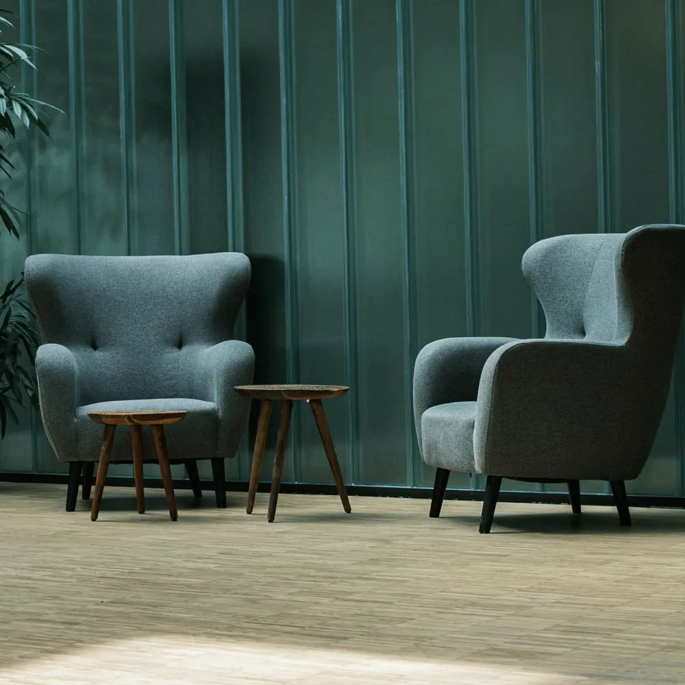 Dve zelene fotelje sa visokim naslonima i dva mala drvena stola u minimalističkom prostoru koji odiše smirenošću.