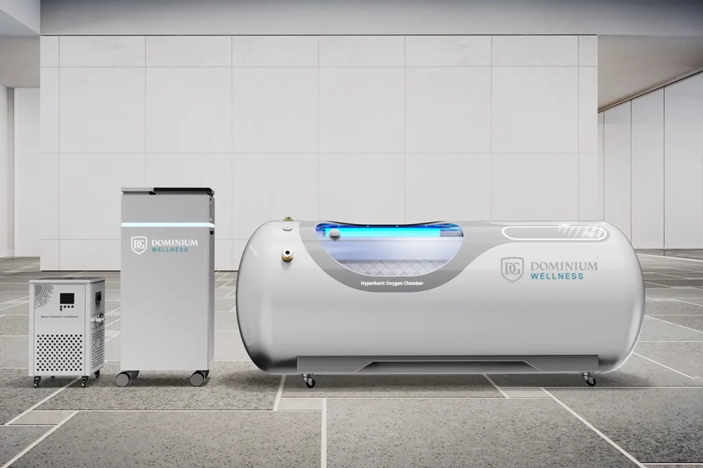 Elegantna, bela hiperbarična komora za kiseonik sa natpisom „Dominium Wellness“ stoji na pločnom podu, okružena sa dve kompaktne naprave, što odražava savremenu medicinsku tehnologiju.